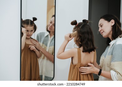 Little girl doing her hair in front of a mirror, face in a reflection. Her mom is watching. - Shutterstock ID 2210284749