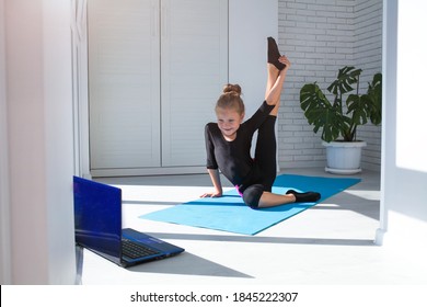 Little Girl Doing Yoga Raised Her Leg Up And Watching Online Lessons On Laptop While Exercising In The Room. A Child Plays Sports During COVID-19