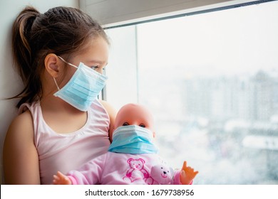 Little Girl With Doll Wearing A Protective Mask. Copy Space. Sadness Kid At Home In Isolation. Sick Child Little Girls Look At The Window With Longing On The Street                                   