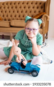 A little girl with Down syndrome in a green dress with glasses to improve vision sits on the floor and plays with toys, in the background is a brown sofa. Help concept for children with special needs.