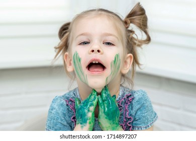 Little girl draws with fingers. Smiling girl touches paint on her cheeks with her palms and leads them down to her neck, self-isolation, coronavirus disease-19, stay home