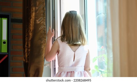 Little Girl In Dress Opened Door And Is Waiting For Guests. Rear View. Waiting For Child Stands Window