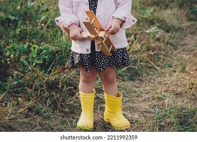 A Little Girl In A Dress And Yellow Rubber Boots Holds A Wooden Airplane, Close-up, No Face. Air Travel, Travel Concept. Children's Games. Retro Style.