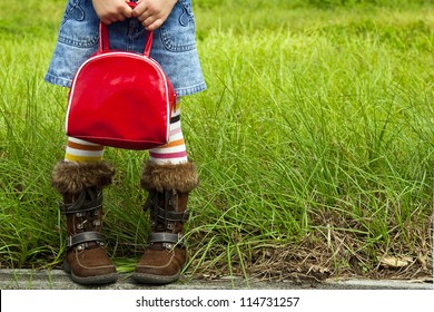 Little girl dressed in striped tights and a denim skirt is standing by a field and holding a red bag