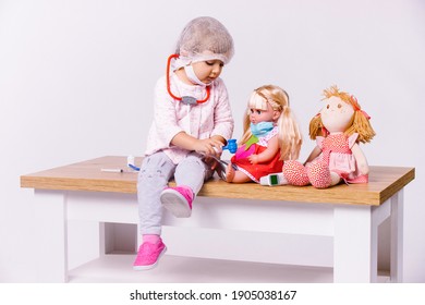 A little girl, dressed in a white medical gown and a medical mask, plays a doctor and treats her toys for various diseases on a white background with side space.