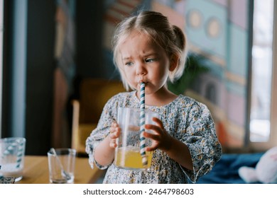 Little girl drinks juice from a glass through a straw while standing at a table in a cafe - Powered by Shutterstock