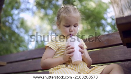 Little girl drinks a milkshake through a straw. Close-up portrait of cute child girl sitting on park bench and drinking milkshake