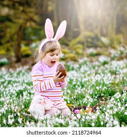 Little Girl With Easter Bunny Ears Making Egg Hunt In Spring Forest On Sunny Day, Outdoors. Cute Happy Child With Lots Of Snowdrop Flowers, Huge Chocolate Egg And Colored Eggs