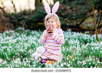 Little Girl With Easter Bunny Ears Making Egg Hunt In Spring Forest On Sunny Day, Outdoors. Cute Happy Child With Lots Of Snowdrop Flowers, Eating Huge Chocolate Egg And Colored Eggs.