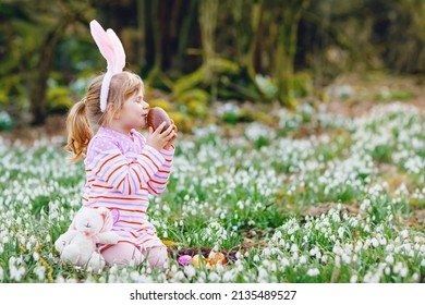 Little Girl With Easter Bunny Ears Making Egg Hunt In Spring Forest On Sunny Day, Outdoors. Cute Happy Child With Lots Of Snowdrop Flowers, Huge Chocolate Egg And Colored Eggs.