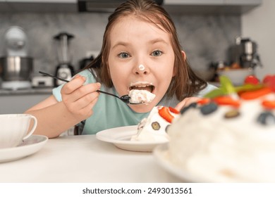 Little girl eating cake while sitting in the kitchen. A pre-teenage girl, stained in cream, eats dessert with a spoon. Selected Focus. High quality photo - Powered by Shutterstock