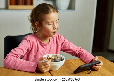 Little Girl Eating Cereal With Milk And Watching Video On Smartphone, Tasty Healthy Food, Slow Motion Of Beautiful Child Having Breakfast At Home, Dependence From Gadget