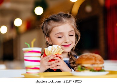 Little girl eating hamburger and French fries in a fast food restaurant. Child having sandwich and potato chips for lunch. Kids eat unhealthy fat food. Grilled fastfood sandwich for children.