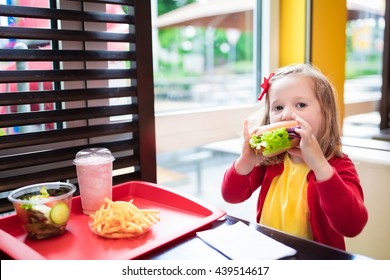 Little Girl Eating Hamburger And French Fries In A Fast Food Restaurant. Child Having Sandwich And Potato Chips For Lunch. Kids Eat Unhealthy Fat Food. Grilled Fastfood Sandwich For Children.