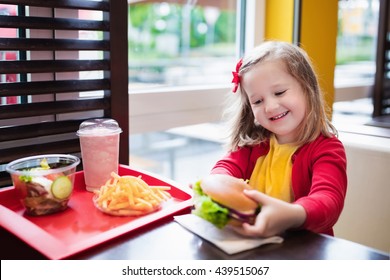 Little Girl Eating Hamburger And French Fries In A Fast Food Restaurant. Child Having Sandwich And Potato Chips For Lunch. Kids Eat Unhealthy Fat Food. Grilled Fastfood Sandwich For Children.