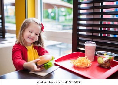 Little Girl Eating Hamburger And French Fries In A Fast Food Restaurant. Child Having Sandwich And Potato Chips For Lunch. Kids Eat Unhealthy Fat Food. Grilled Fastfood Sandwich For Children.