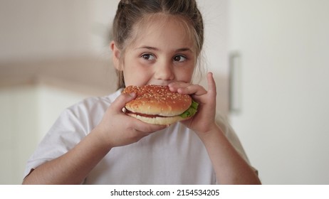 Little Girl Eating A Hamburger. Unhealthy Fast Food Proper Nutrition Concept. Child Greedily With Pleasure Lifestyle Bites A Big Burger In The Kitchen At Home. Kid Eats Fast Food Close-up