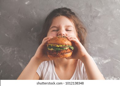 The Little Girl Is Eating A Healthy Baked Sweet Potato Burger With A Whole Grains Bun, Guacamole, Vegan Mayonnaise And Vegetables. Child Vegan Concept