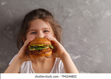 The Little Girl Is Eating A Healthy Baked Sweet Potato Burger With A Whole Grains Bun, Guacamole, Vegan Mayonnaise And Vegetables. Child Vegan Concept