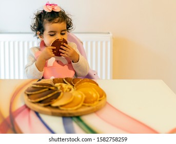 A Little Girl Eating Pancakes For Breakfast. Wooden Serving Plate. Child Wearing Bib. Baby Girl Smelling Food