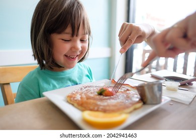 Little girl enjoying delicious pancakes with maple syrup and a strawberry for breakfast on a sunny summer day in Hawaii. Mother gives a helping hand.