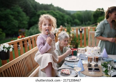 Little Girl Enjoying Eating Birthday Cake During Multi Generation Family Celebration Outside On Patio.