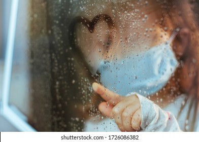 Little Girl In Face Mask Drawing Heart Symbol On The Window Glass With Rain Drops. Selective Focus On The Heart. Social Isolation Stay At Home During Pandemic COVID-19 Concept.