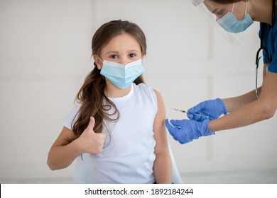 Little girl in face mask gesturing thumb up while receiving covid-19 vaccine injection at health centre. Cute child approving of coronavirus immunization, being protected against global virus