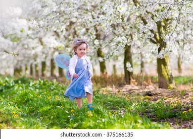 Little Girl In Fairy Costume With Wings, Flower Crown And Magic Wand Playing In Blooming Apple Tree Garden On Easter Egg Hunt. Kid Watching Cherry Blossom In The Garden. Child In Spring Fruit Orchard.