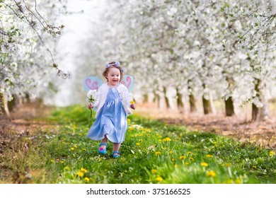 Little Girl In Fairy Costume With Wings, Flower Crown And Magic Wand Playing In Blooming Apple Tree Garden On Easter Egg Hunt. Kid Watching Cherry Blossom In The Garden. Child In Spring Fruit Orchard.