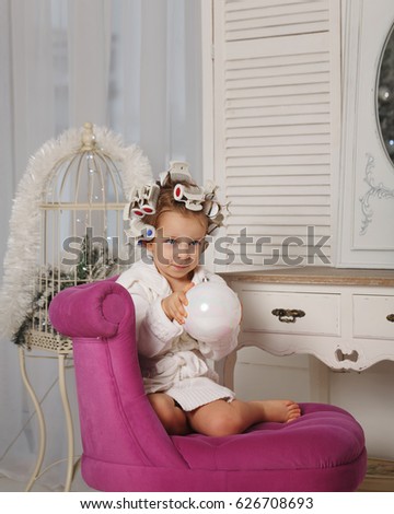 Little girl fashionista. The girl in the curler and robe sits on the chair barefoot. Little coquette. Human emotions.