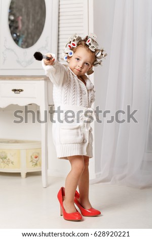 Little girl fashionista. A girl in a curler, a robe and red high-heeled shoes is holding a makeup brush. Little coquette posing. Human emotions. As an adult.