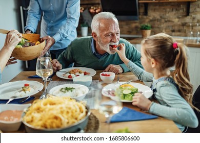 Little Girl Feeding Her Grandfather While Having Family Lunch In Dining Room. Focus Is On Senior Man. 