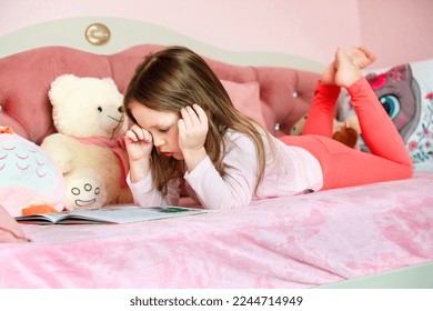 una niña de cinco años de largo cabello yace en la acogedora cama de sus hijos con juguetes y lee un libro, frota los ojos y quiere dormir