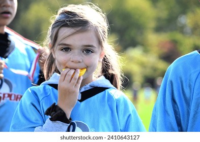 Little girl Flag Football player looking straight ahead, holding her yellow mouth-guard in her mouth with her fingers. Chilly, bright fall morning. Green trees and grass background.