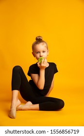 Little Girl Full Body In A Swimsuit, Bites A Green Apple, Sitting On A Yellow Background During A Break In Training