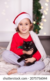 Little Girl In Funny Santa Hat And Christmas Pajamas With Favorite Pet Black Cat