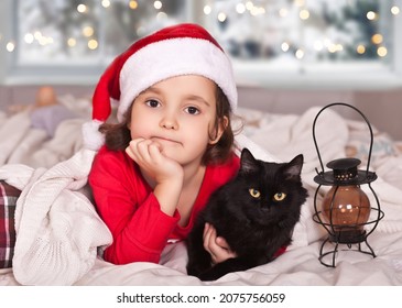 Little Girl In Funny Santa Hat And Christmas Pajamas With Favorite Pet Black Cat