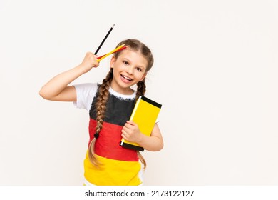 A little girl with a German flag on her T-shirt holds a book and pencils on a white isolated background. German language courses for schoolchildren.