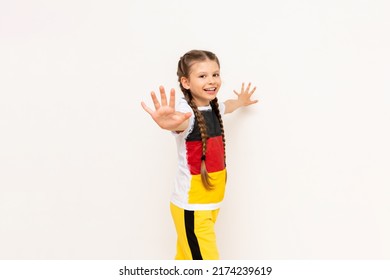 A little girl with a German flag on her T-shirt, with long hair braided into pigtails, stretches her palm towards you and smiles broadly against a white isolated background. Language courses. 