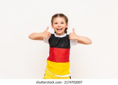 A little girl with a German flag on a T-shirt, with long hair in pigtails, gives a thumbs up and smiles broadly on a white isolated background. Language courses.