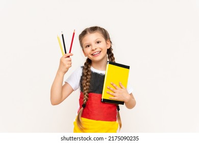 A little girl with a German flag on her T-shirt holds a book and pencils on a white isolated background. German language courses for schoolchildren.
