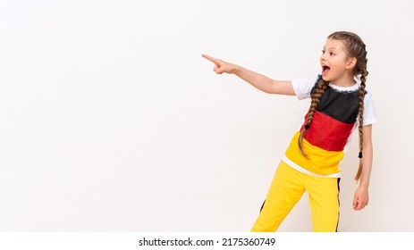 A little girl with a German flag on a T-shirt, with long hair in pigtails, points up with her index finger sideways at your advertisement on a white isolated background. Language courses. Copy space.