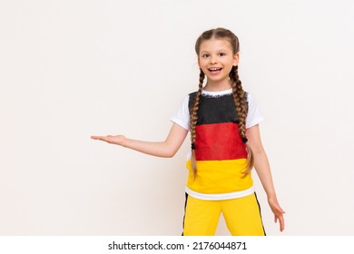 A little girl with a German flag on a T-shirt, with long hair in pigtails, holds your advertisement on a white isolated background on her palms. Language courses. Copy space.