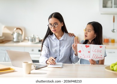 Little Girl Giving Greeting Card To Her Busy Mother, Irritated Mom Not Paying Attention, Sitting In Kitchen Interior. Working Woman Having No Time For Her Kid