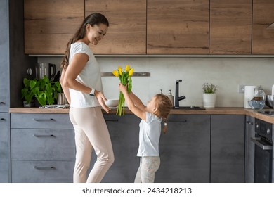 Little girl giving yellow tulips to her mum, while woman cooking on the kitchen. Mothers day, birthday or International Women's Day 8 march concept. High quality photo - Powered by Shutterstock