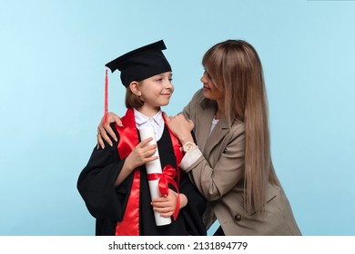 Little Girl Graduate Celebrating Graduation. Child Wearing Graduation Cap And Ceremony Robe Holding Certificate. Mom Hugs And Congratulations Daughter On Graduation. Successfully Complete Course Study