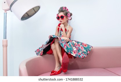 Little girl with hair curlers and red sunglasses sitting on couch, near the old style hair dryer, Kid's fashion - Powered by Shutterstock