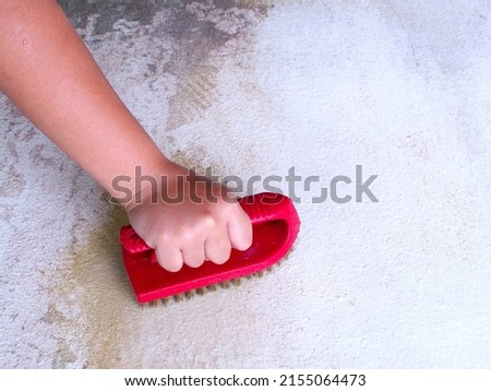 little girl hand uses red plastic brass wire brush scrubbing mud stain on white concrete floor, kid helping chore with cleaning tool washing dirt cement floor, closeup top view with copy space