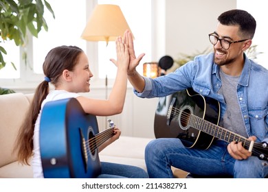 little girl and handsome father play guitar sitting on couch at home, give high five. father's day. care and education of children. side view on nice positive adult man and child girl in casual wear